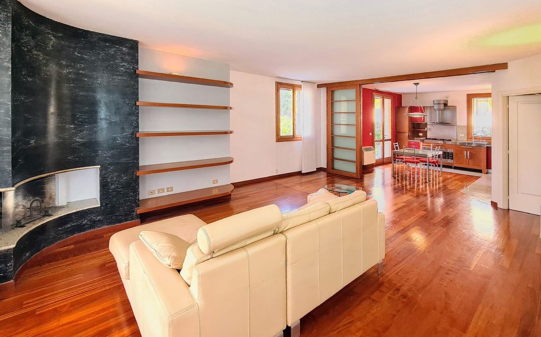 Soggiorno della villa con camino in pietra scura e parquet, aperto sulla cucina